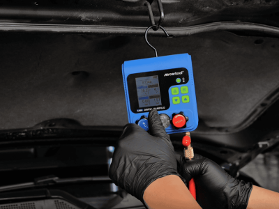 Digital refrigerant manifold gauge in use for auto AC system testing and servicing, with a technician wearing gloves.